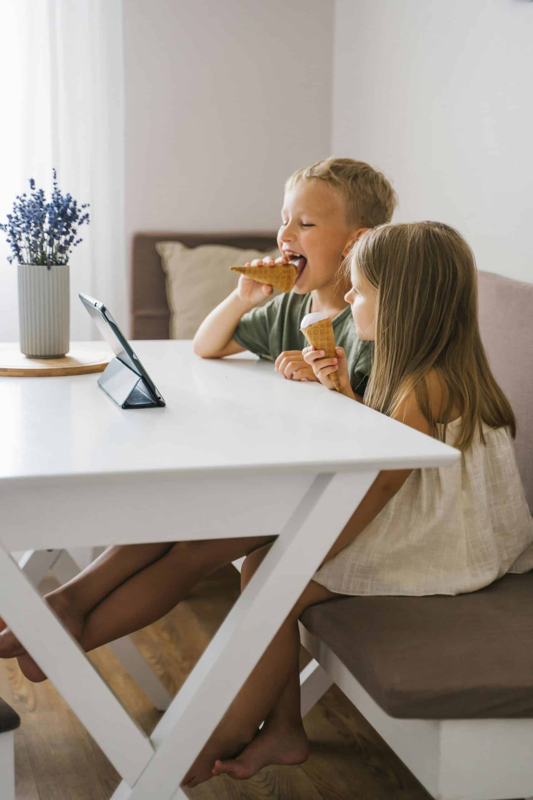 Two children enjoying ice cream cones indoors while watching a tablet together in a cozy setting.