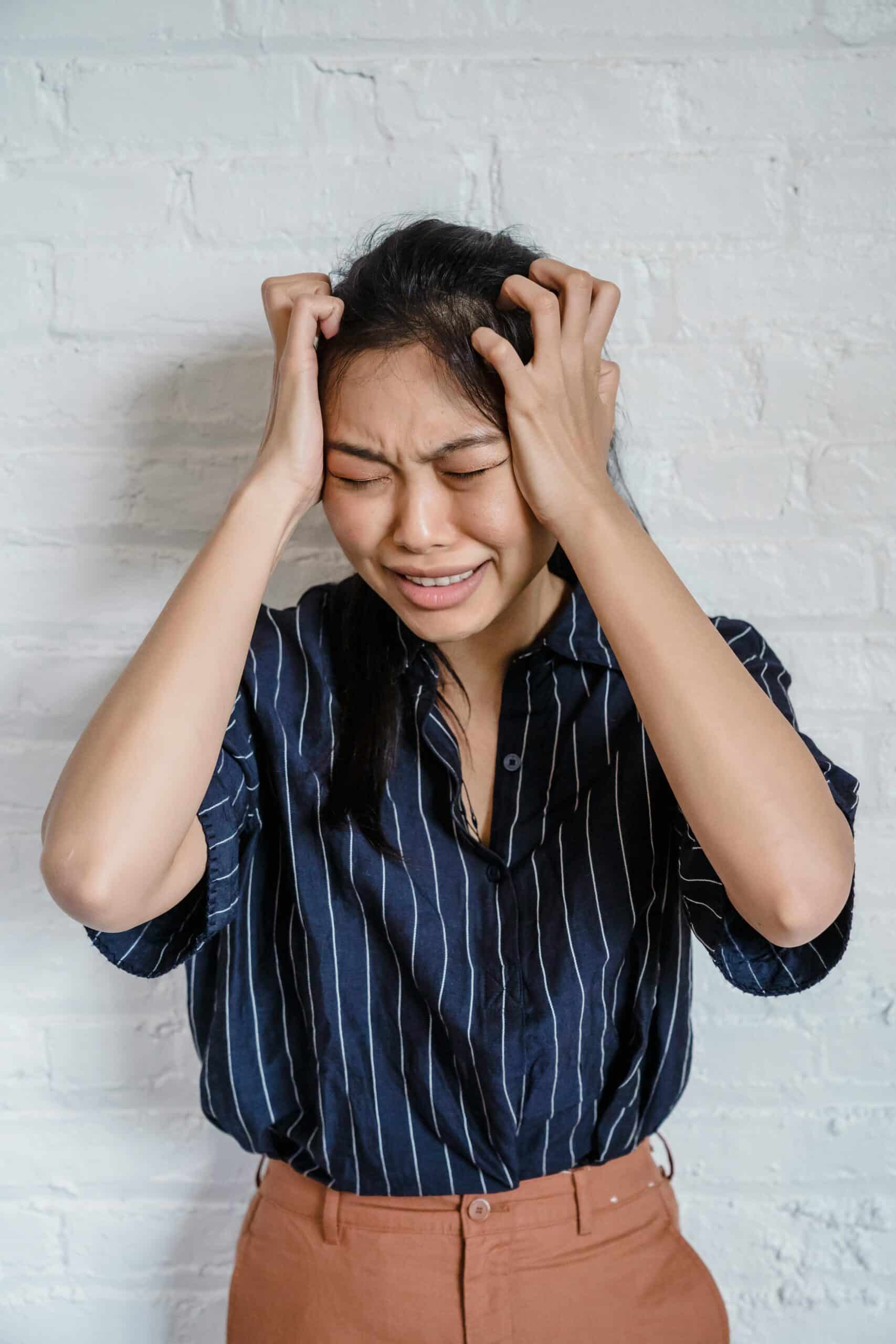 Asian woman with eyes closed, feeling stressed and experiencing a headache against a white brick wall.